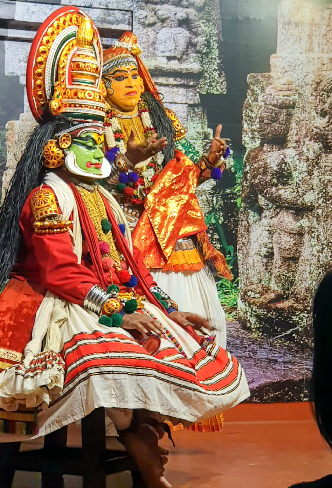 Two performers in traditional Kathakali attire and makeup, one seated and one gesturing with hands, with a backdrop of ancient temple carvings