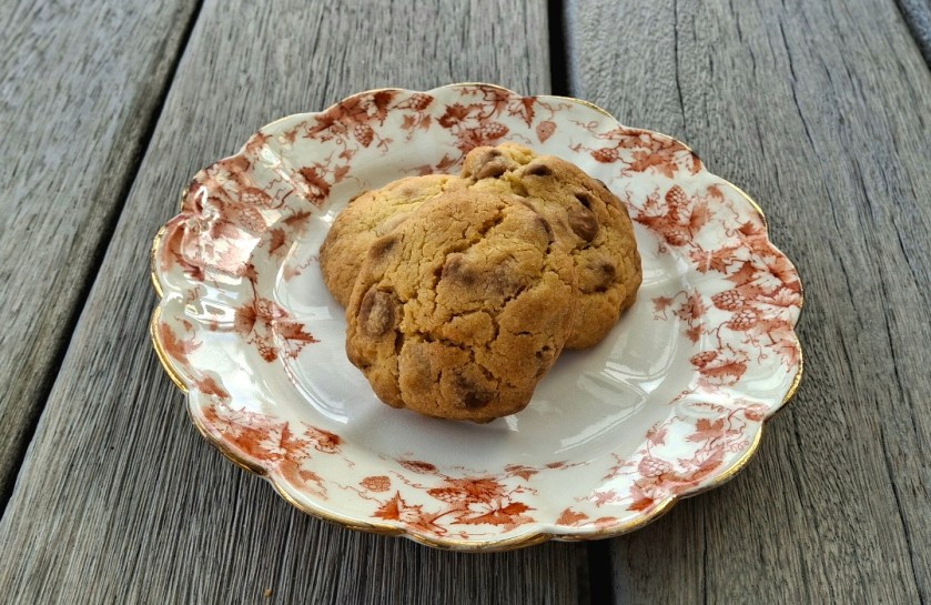 Three chocolate chip cookies on a decorative white plate with red floral patterns on a wooden surface