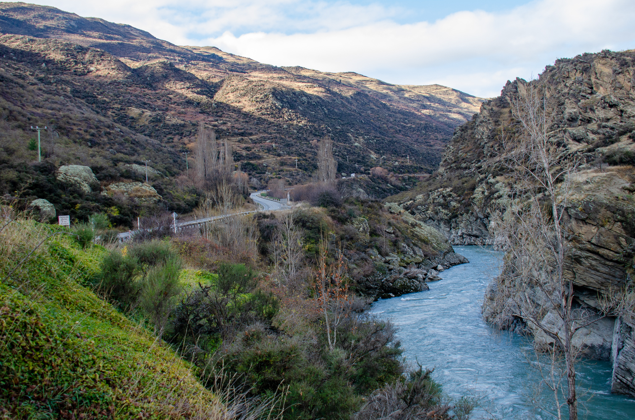 Winding road running along a river through rocky hills with sparse vegetation and leafless trees under a partly cloudy sky