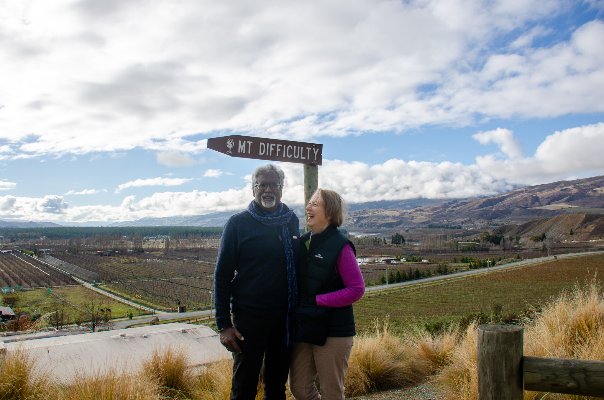 Two people standing and smiling near a sign that says MT DIFFICULTY with a vineyard and mountains in the background under a cloudy sky
